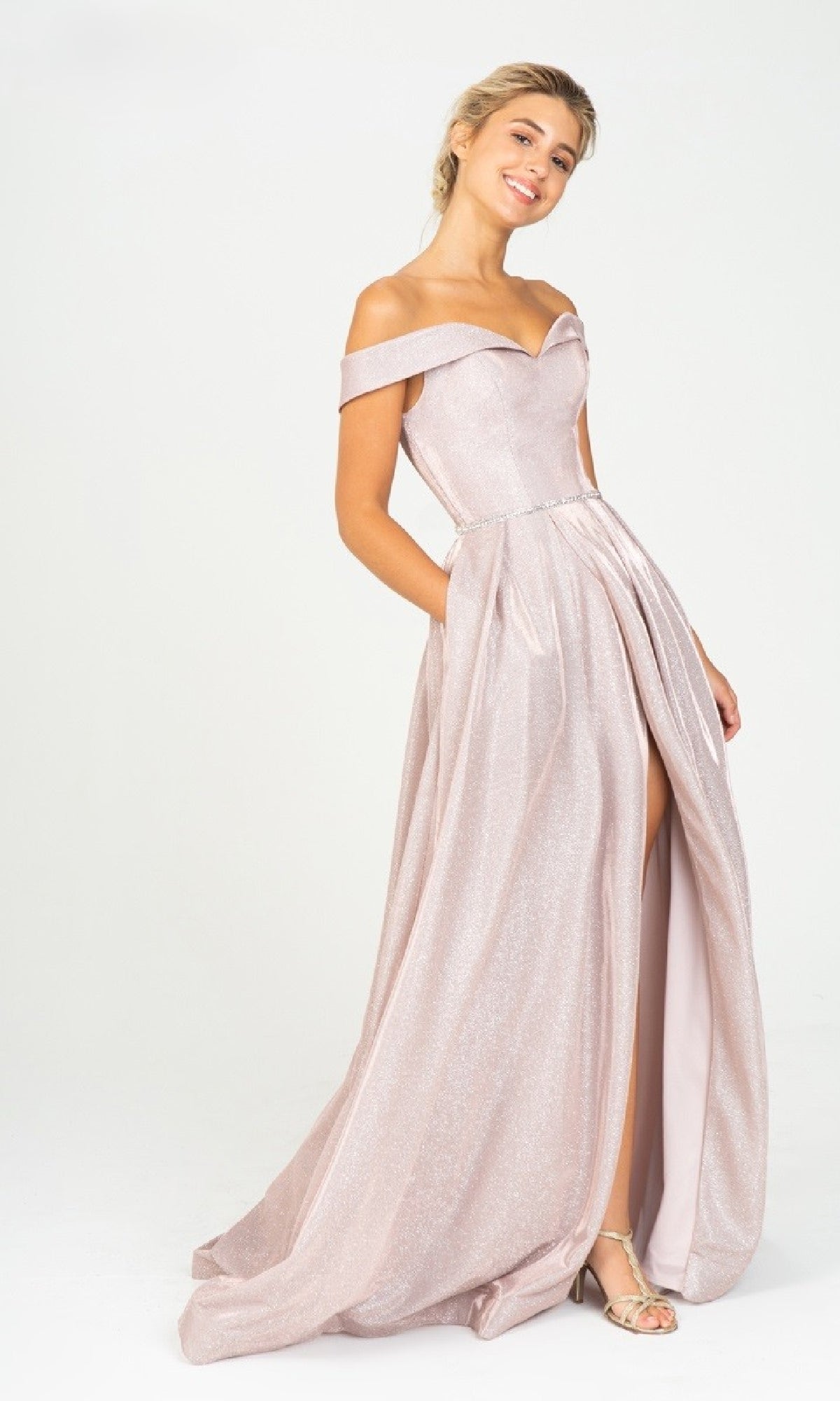 A woman stands smiling against a plain background, wearing the Eureka 9808 Off-Shoulder Long Glitter Prom Dress in light pink, featuring a floor-length design and thigh-high slit.